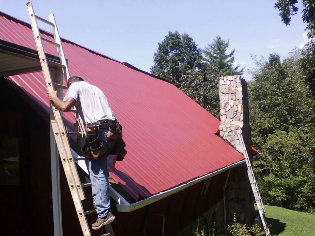 Roofer on a ladder beside a red metal roof and stone chimney — Soffit & Fascia Repair by Roofing Brothers in Varnville